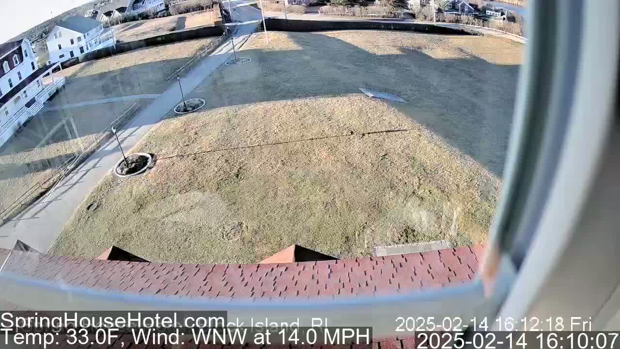 A high-angle view shows a brown, grassy area with a paved walkway, some landscaping, and buildings in the background on a sunny day with a WNW wind at 14 mph and a temperature of 33°F.