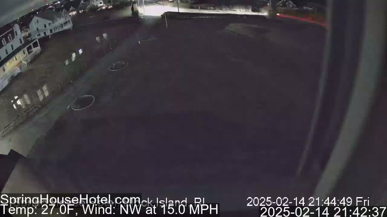 A nighttime, outdoor view of a mostly empty parking lot next to buildings, with a northwest wind at 15 mph and a temperature of 27 degrees Fahrenheit.