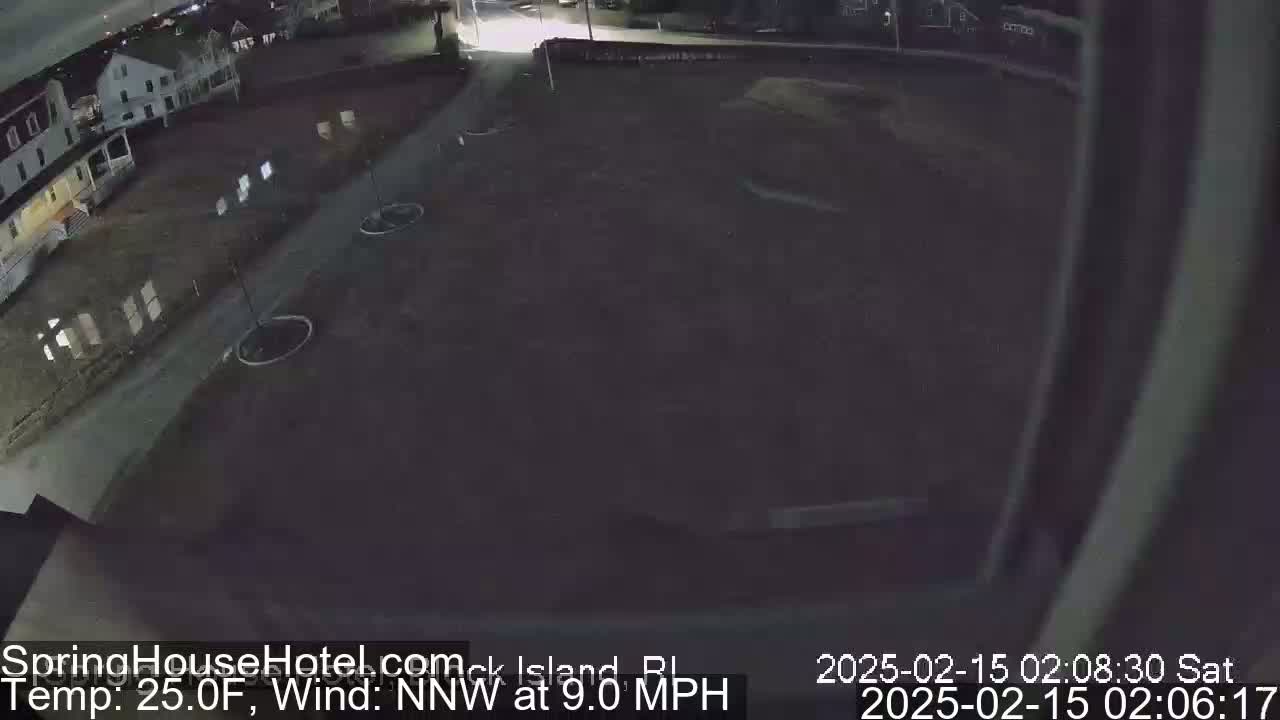 A nighttime, outdoor view of a mostly empty paved area near some houses, under a dark sky with a northwest wind at 9 mph and a temperature of 25 degrees Fahrenheit.