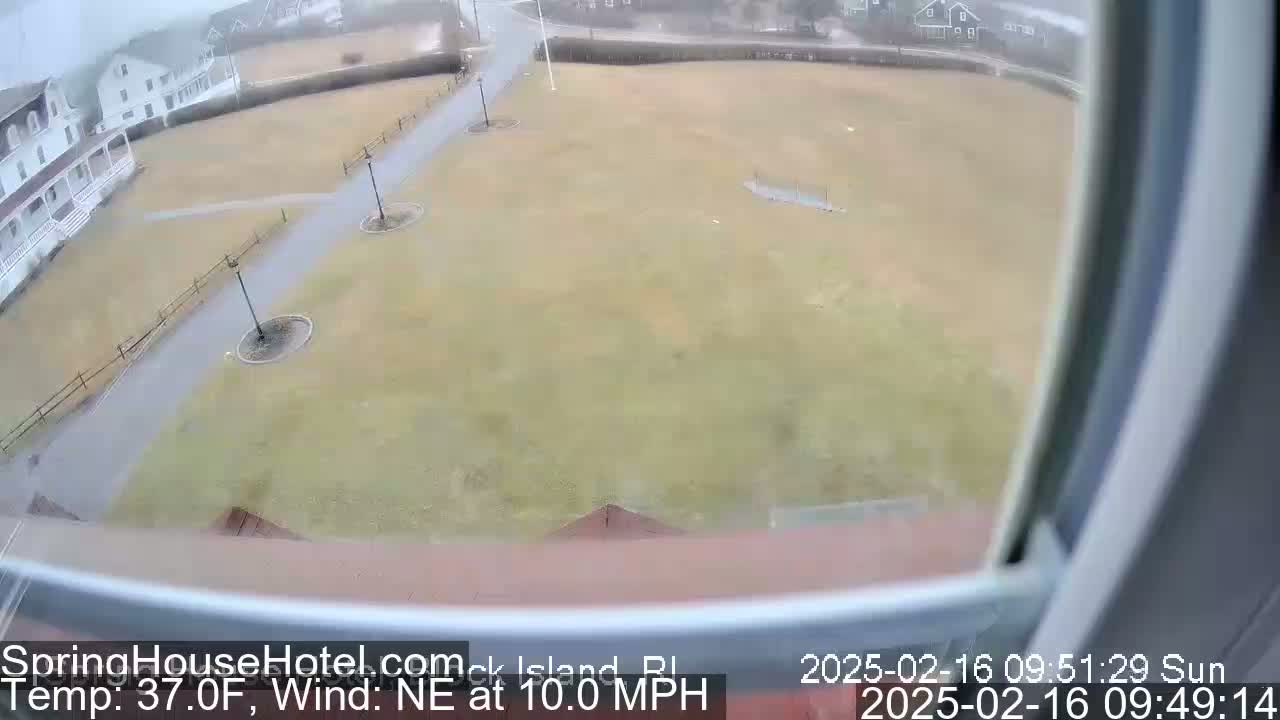 A view from a window shows a grassy area with walkways, light posts, and buildings in the background under a gray, overcast sky.