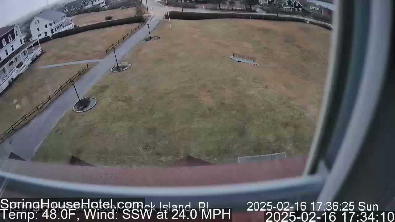 A view from a window shows a large, brown grassy area with paved walkways and a few buildings in the background, under overcast skies with a strong south-southwesterly wind.
