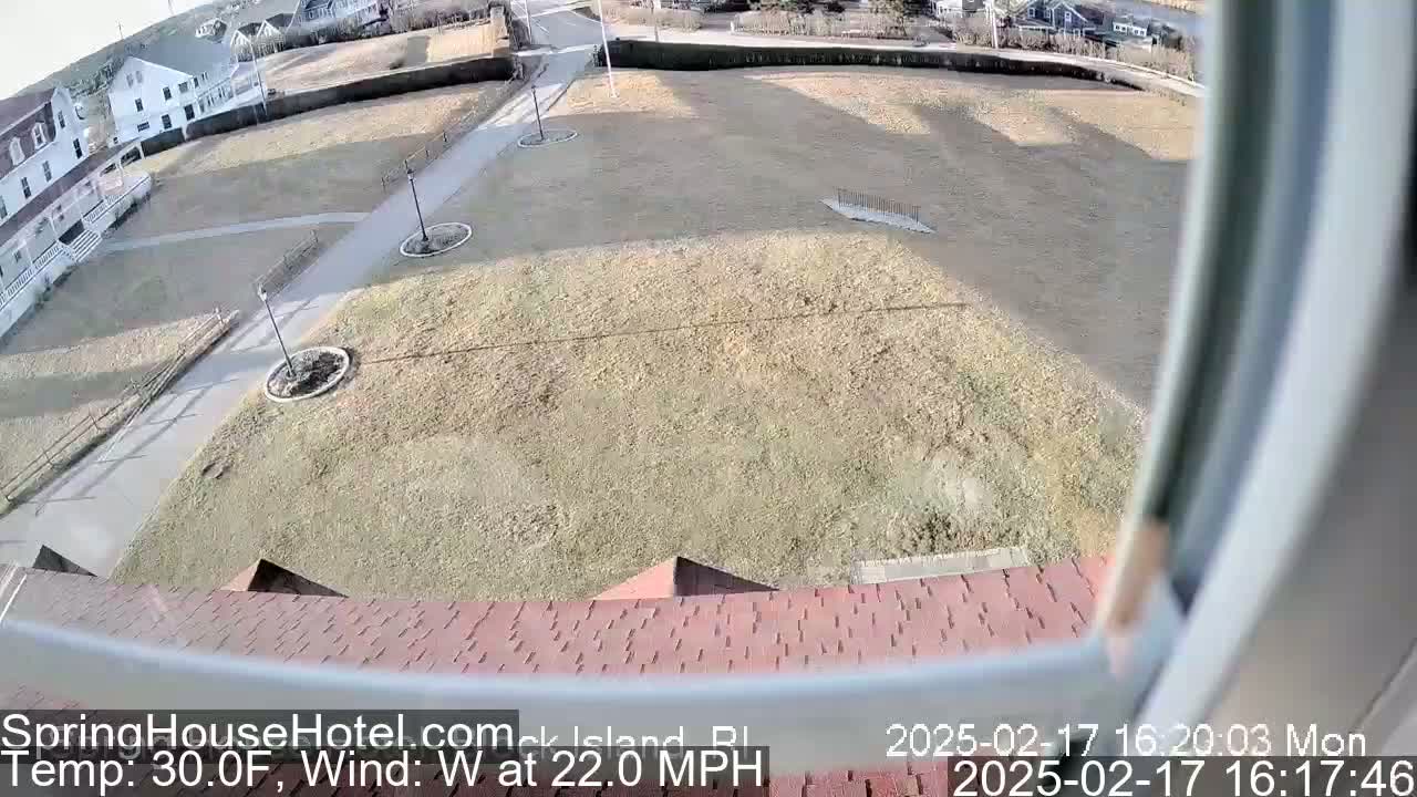 An elevated view of a grassy area with paved pathways, buildings, and a reddish-brown roof in the foreground, on a windy, 30°F day.