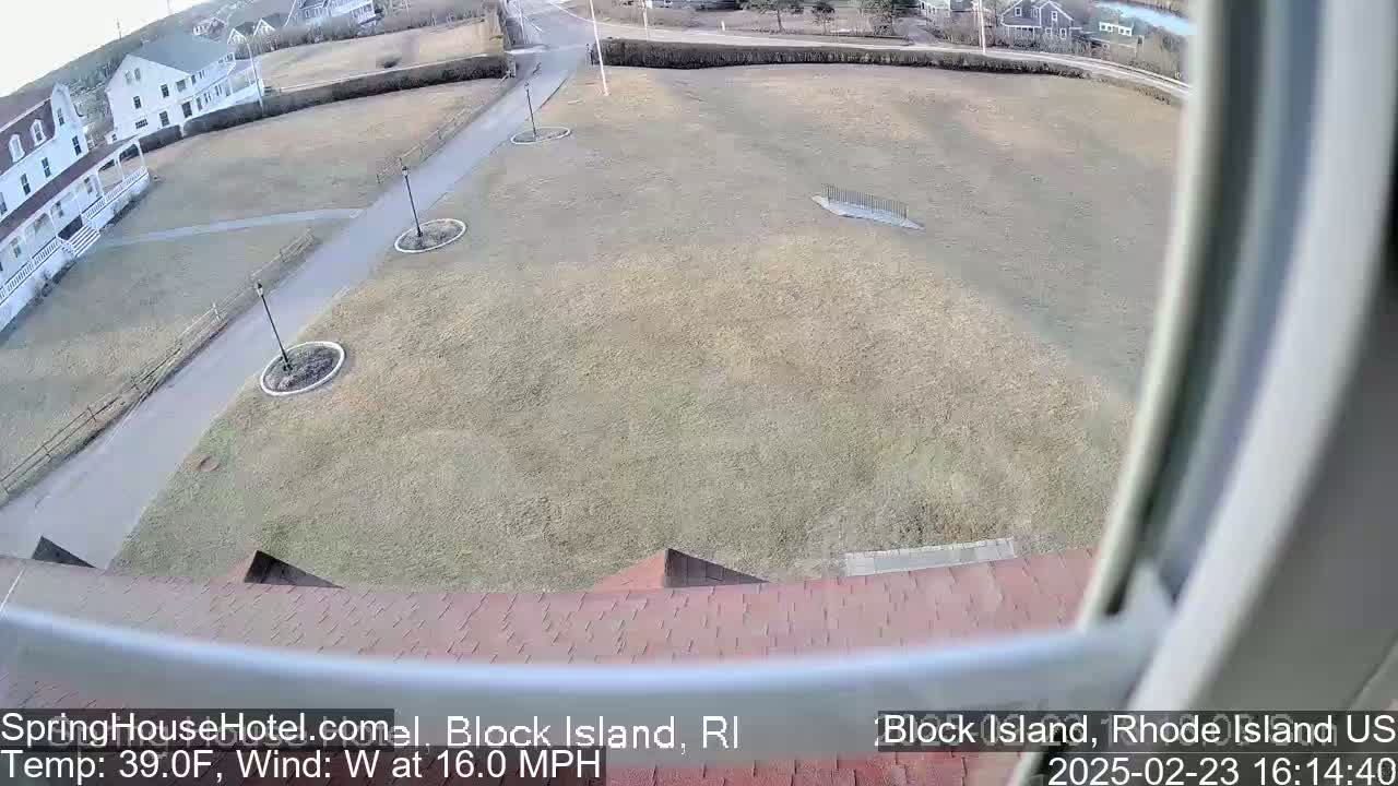 A high-angle view shows a large, brown grassy area with a paved walkway and buildings in the background, under a mostly clear sky with a 16 mph westerly wind and a temperature of 39°F.