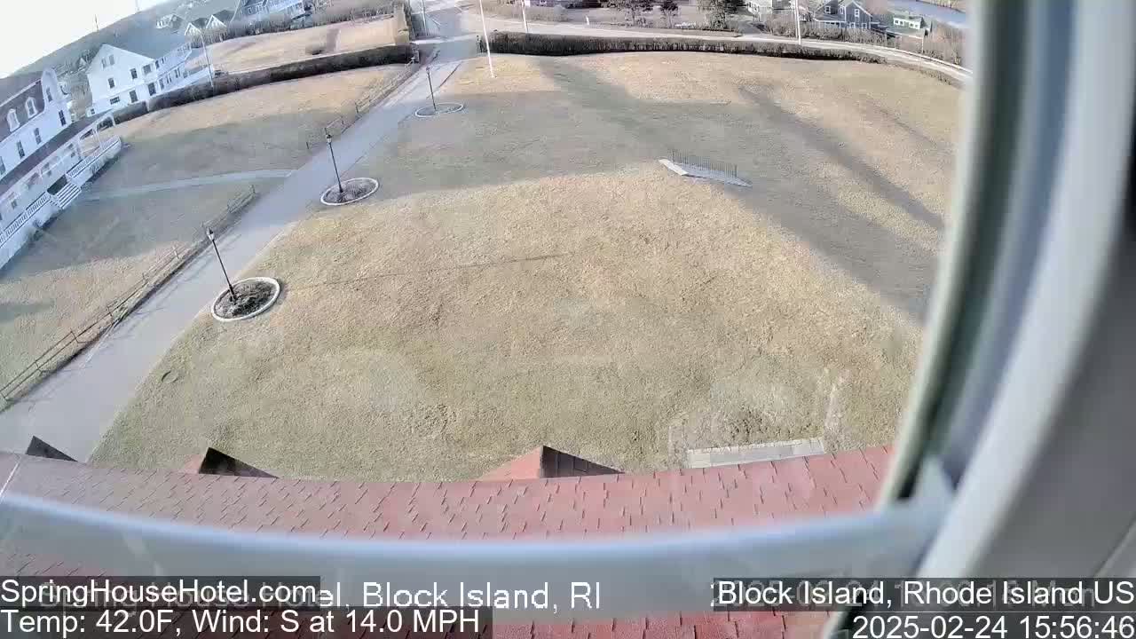 A high-angle view from a window shows a large, mostly barren grassy area with paved walkways, some landscaping, and a building in the background, under sunny conditions with a south wind of 14 mph.