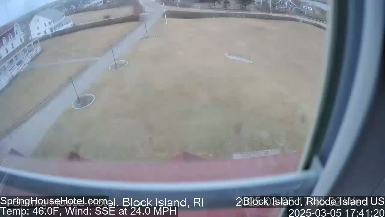 A view from a window shows a grassy area with pathways and lampposts, under overcast skies with a strong wind.