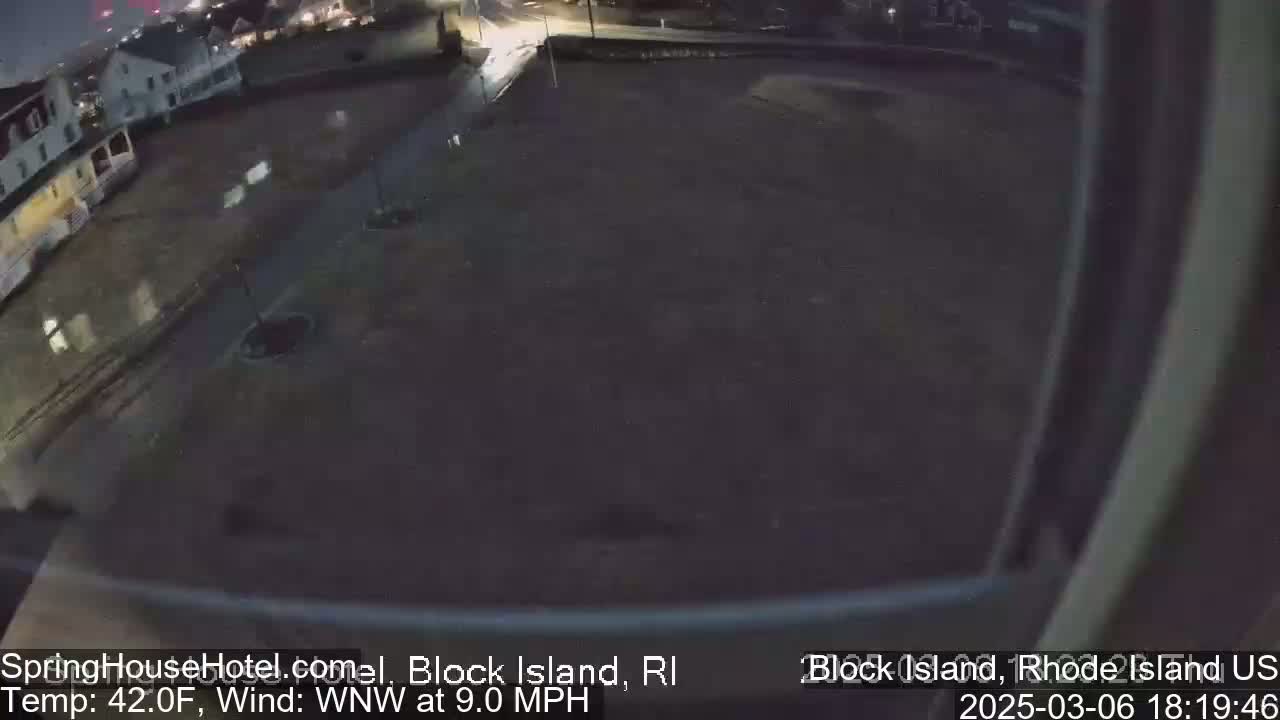 A wet, dark nighttime scene shows a mostly empty paved area next to buildings, with a 9 mph wind.