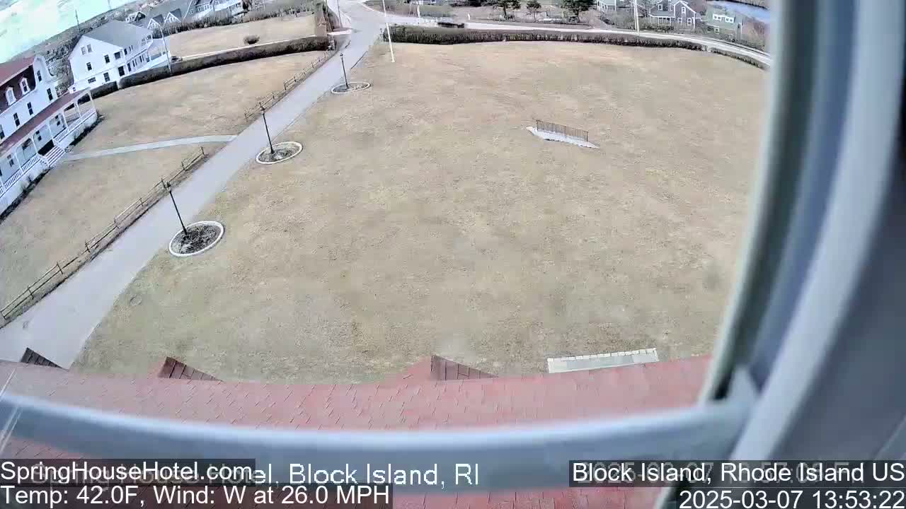 A high-angle view shows a large, brown grassy area with a paved walkway and buildings in the background, under windy, 42°F weather.