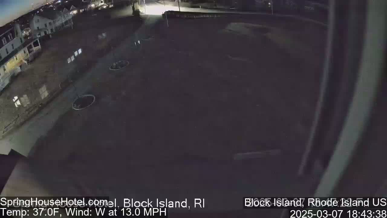 A nighttime, overhead view of a mostly empty parking lot next to a residential street with a few buildings visible in the distance, under a dark sky with a westerly wind.