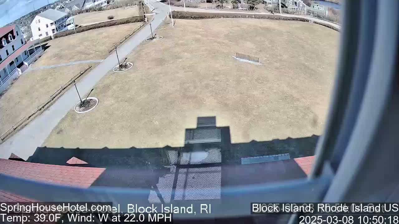 A mostly sunny view from a window shows a large, flat grassy area with a paved walkway, some landscaping, and buildings in the background, with a 39°F temperature and a 22 mph west wind.