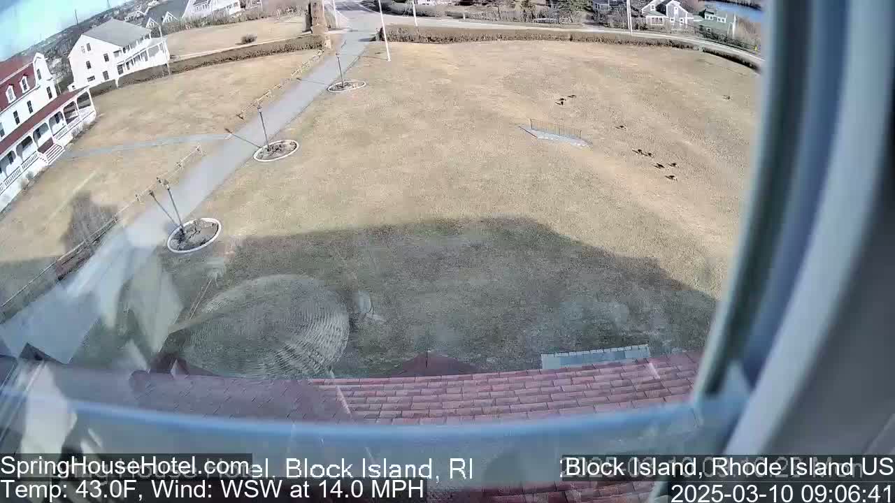 A view from a window shows a brown, grassy field with a few birds and a paved pathway leading to a white building, under sunny conditions with a southwest wind.