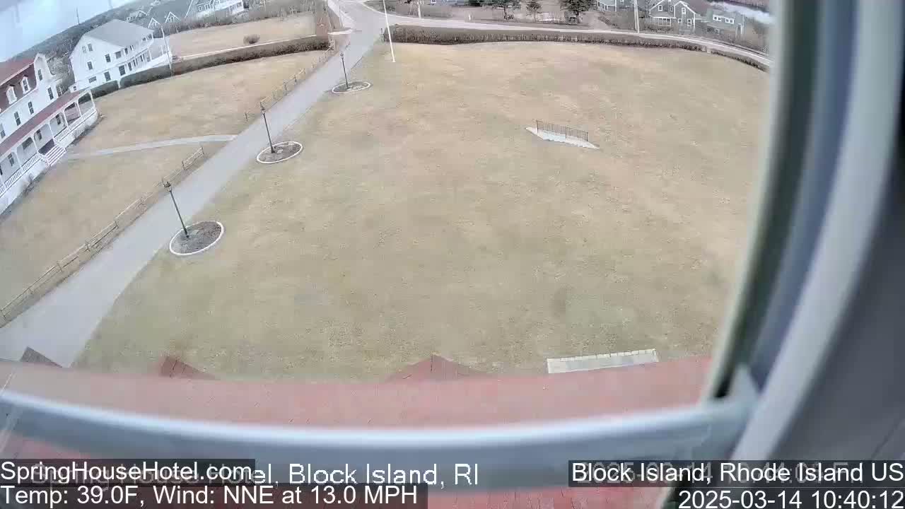 A large, mostly brown grassy area with a paved walkway and a building in the background, viewed from a window on a slightly windy, 39°F day.