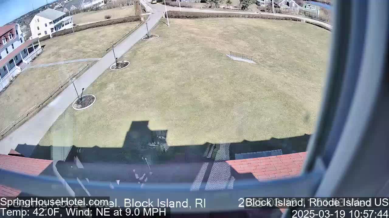 A view from a window shows a sunny, grassy area with a paved walkway and several buildings in the background, under a northeast wind of 9 mph and a temperature of 42°F.