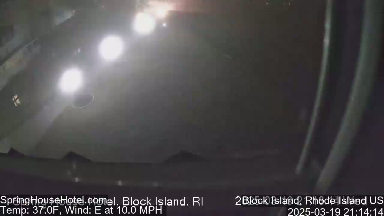 A hazy nighttime scene shows a dark, empty parking lot illuminated by the headlights of several distant vehicles, with a 10 mph east wind and a temperature of 37°F.