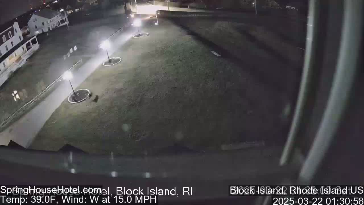 A nighttime, overhead view of a grassy area with a paved pathway, several light posts, and a building in the background, under windy conditions.