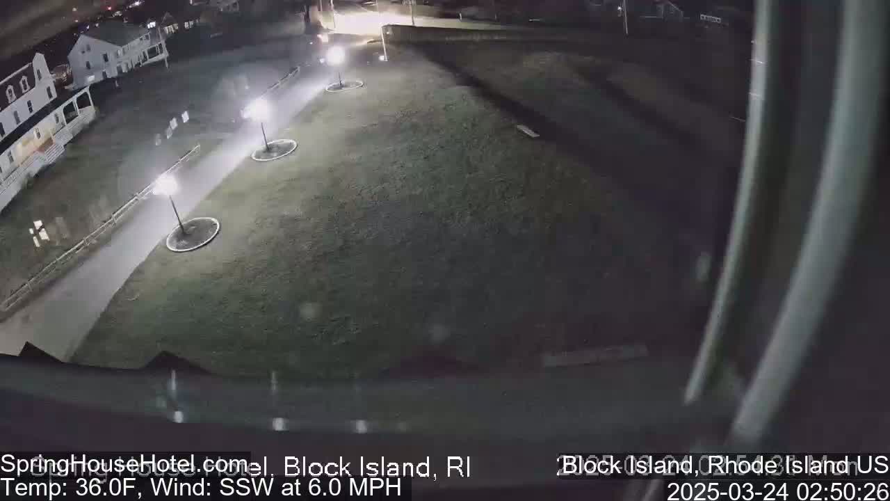 A nighttime, aerial view shows a grassy area with three lit lampposts, a paved path, and a building in the background under a slightly snowy, windy sky.