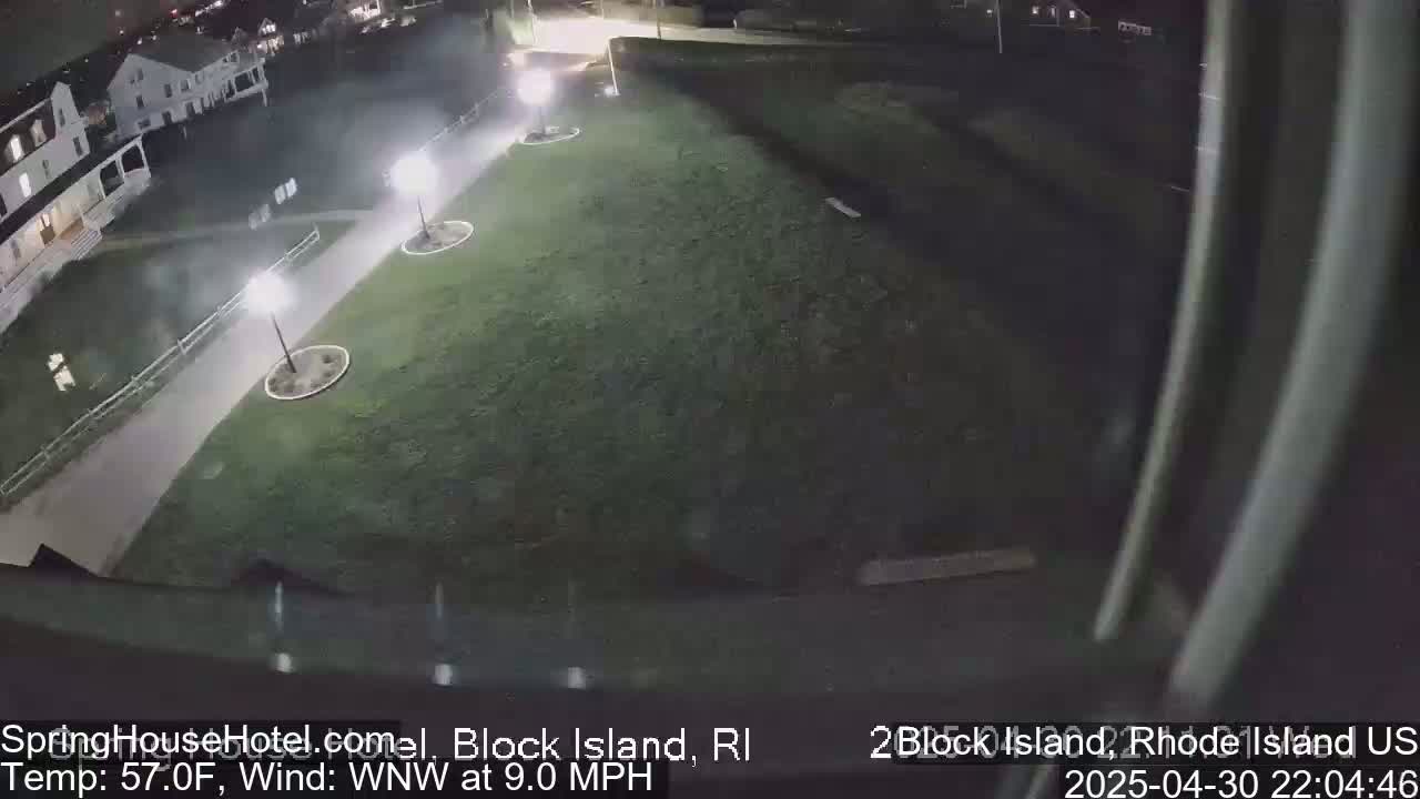 A nighttime, high-angle view shows a grassy area with a paved walkway, several lampposts, and a building in the background under a clear sky with a wind of 9 mph.