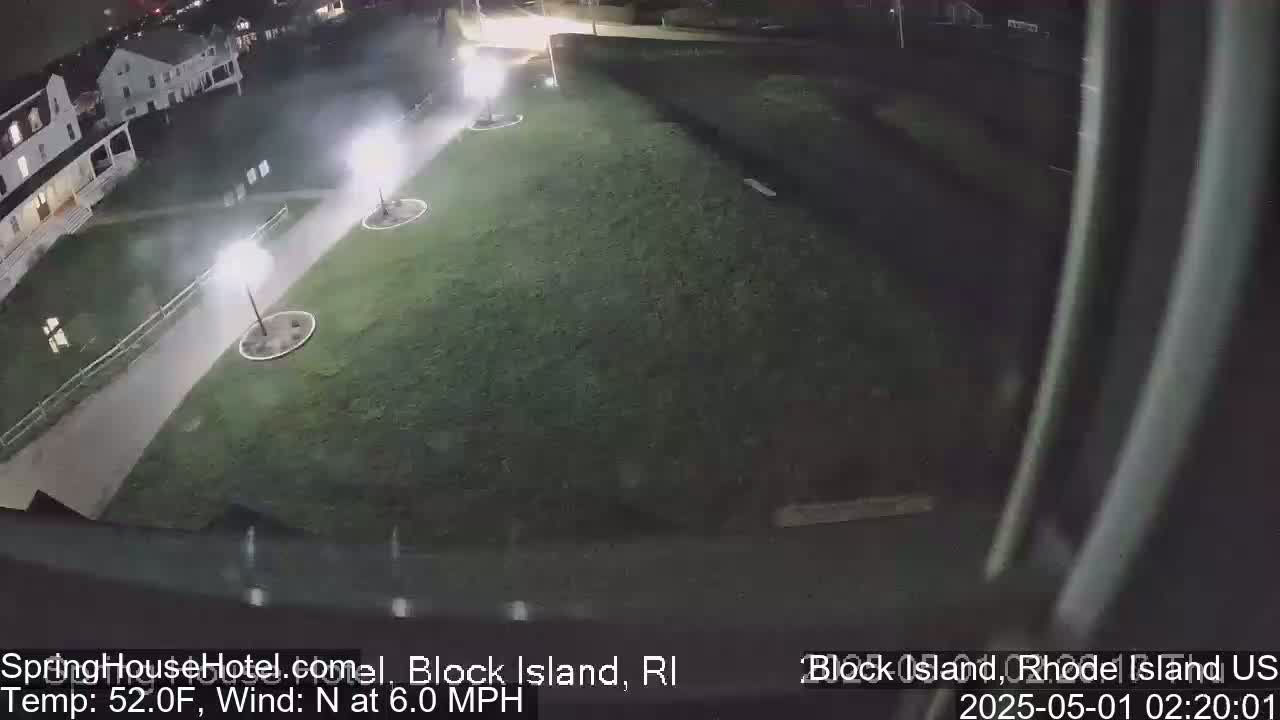 A nighttime, overhead view of a grassy area with several lampposts illuminating a walkway next to a building under a clear sky with a 6 mph north wind.