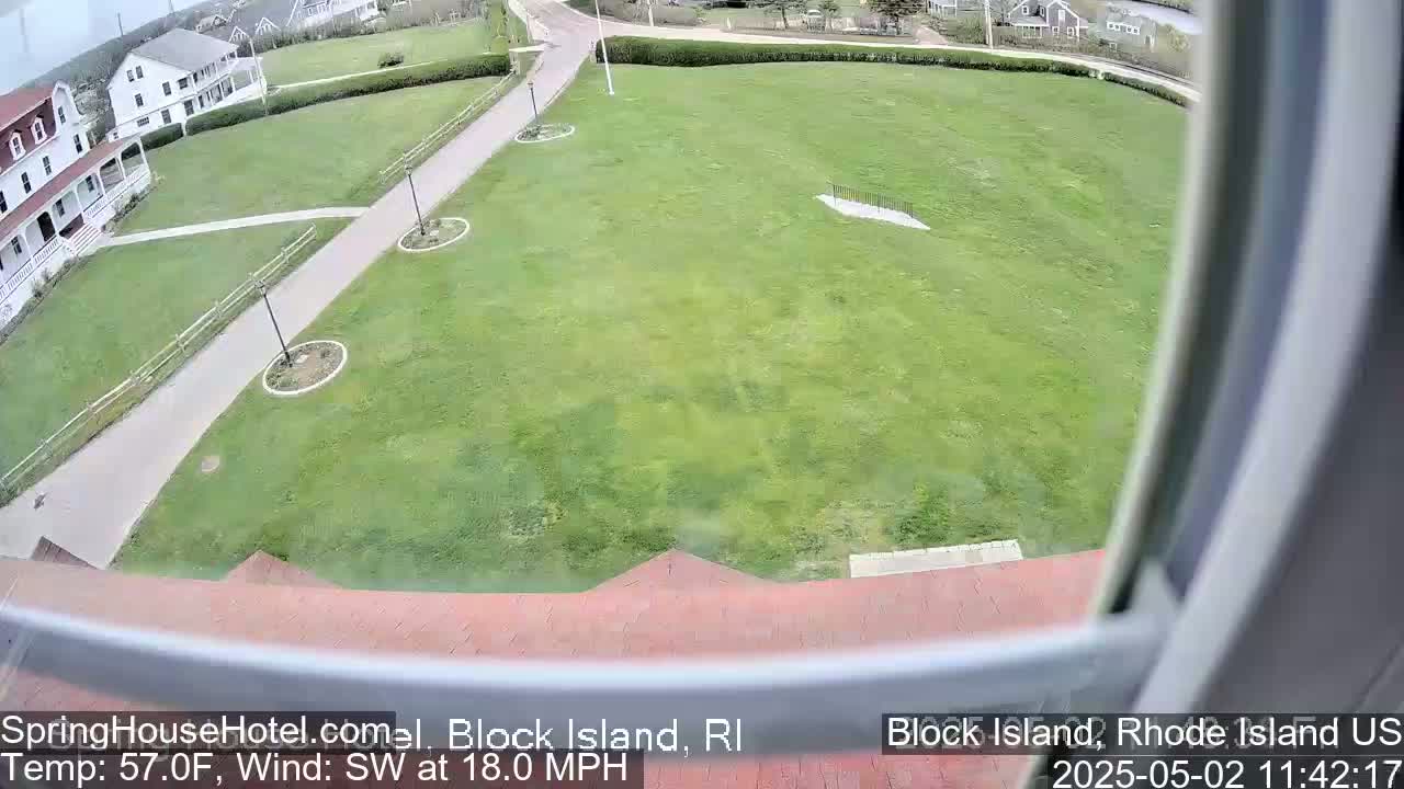 A view from a window shows a large grassy area with walkways, landscaping, and a white building in the background, all under a partly cloudy sky with a southwest wind of 18 mph.