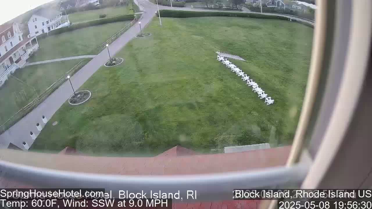 A high-angle view shows a grassy lawn with a line of white chairs, a paved walkway, and a building in the background, all under a partly cloudy sky with a south-southwesterly wind at 9 mph and a temperature of 60 degrees Fahrenheit.