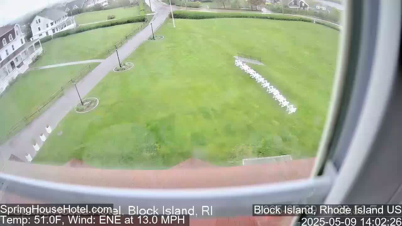 A view from a window shows a green lawn with a row of white chairs, a building, and a paved pathway, all under overcast skies.