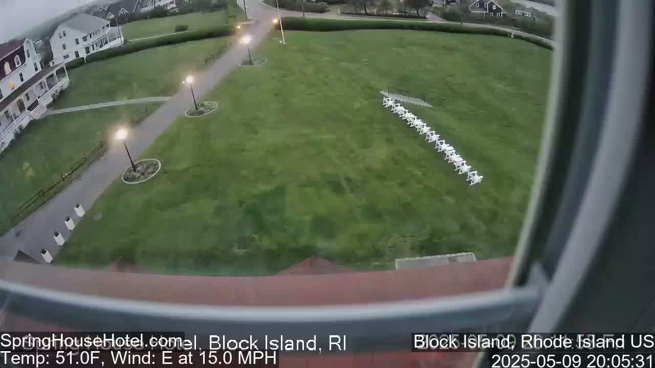 A grassy lawn with a line of white chairs is visible from a window, with a building and a paved pathway in the background, under an overcast sky with a 15 mph east wind and 51°F temperature.