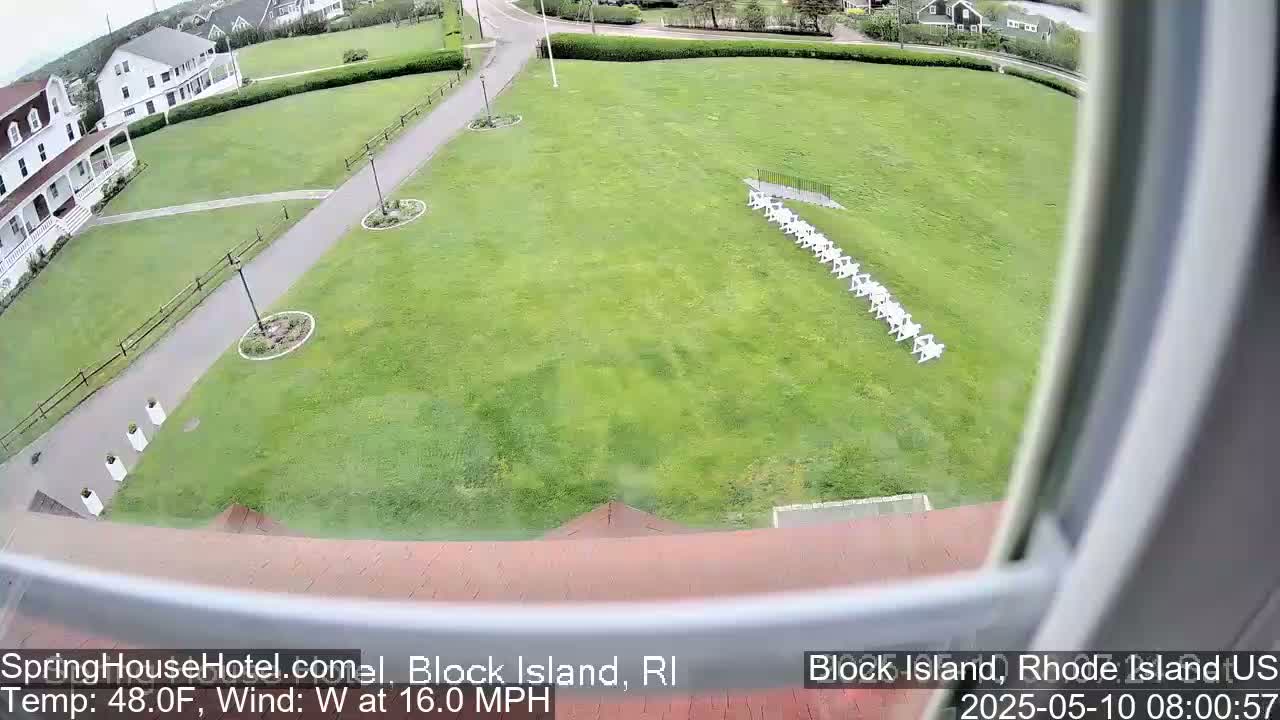 A view from a window shows a grassy lawn with a row of white chairs, a building, and a paved path, all under a cloudy sky with a 16 mph westerly wind and a temperature of 48°F.