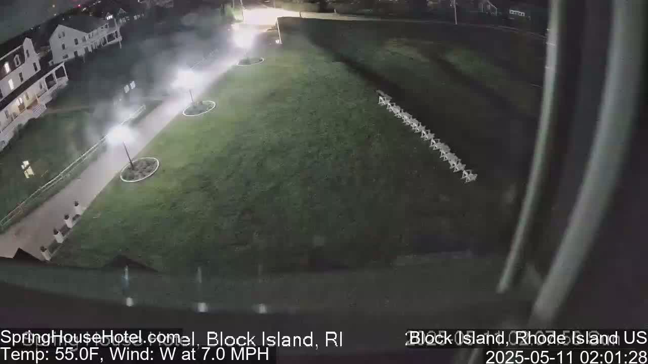 A nighttime, slightly hazy view shows a grassy area with a row of chairs, a paved walkway with lights, and a building in the background, under a 55°F temperature with a 7 mph west wind.