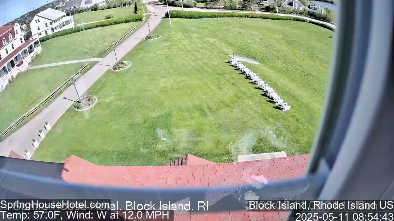 A large grassy area with a row of white chairs, a walkway, and a white building visible in the background on a sunny day with a 12 mph westerly wind.