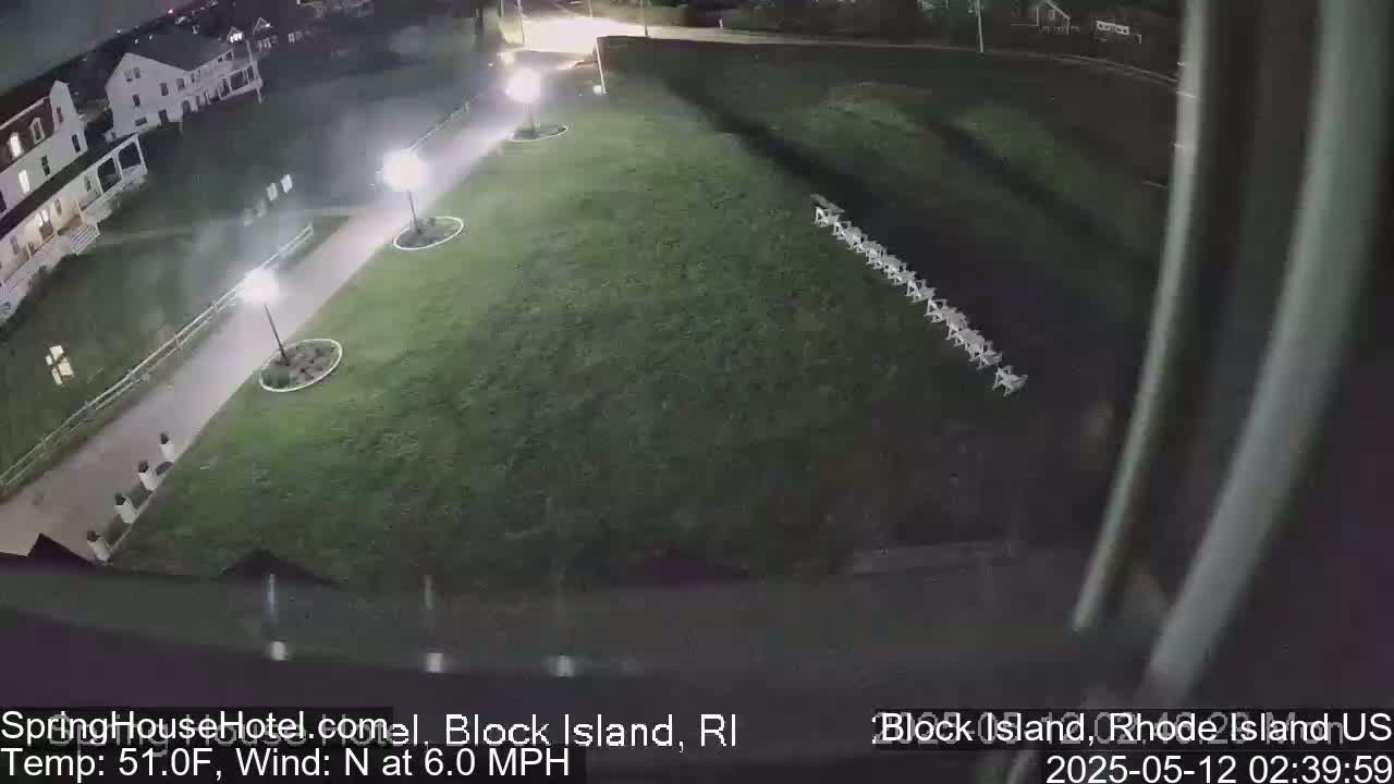 A nighttime, overhead view shows a grassy area with a row of chairs, a paved walkway with lights, and a building in the background, under a 51°F temperature with a 6.0 MPH north wind.