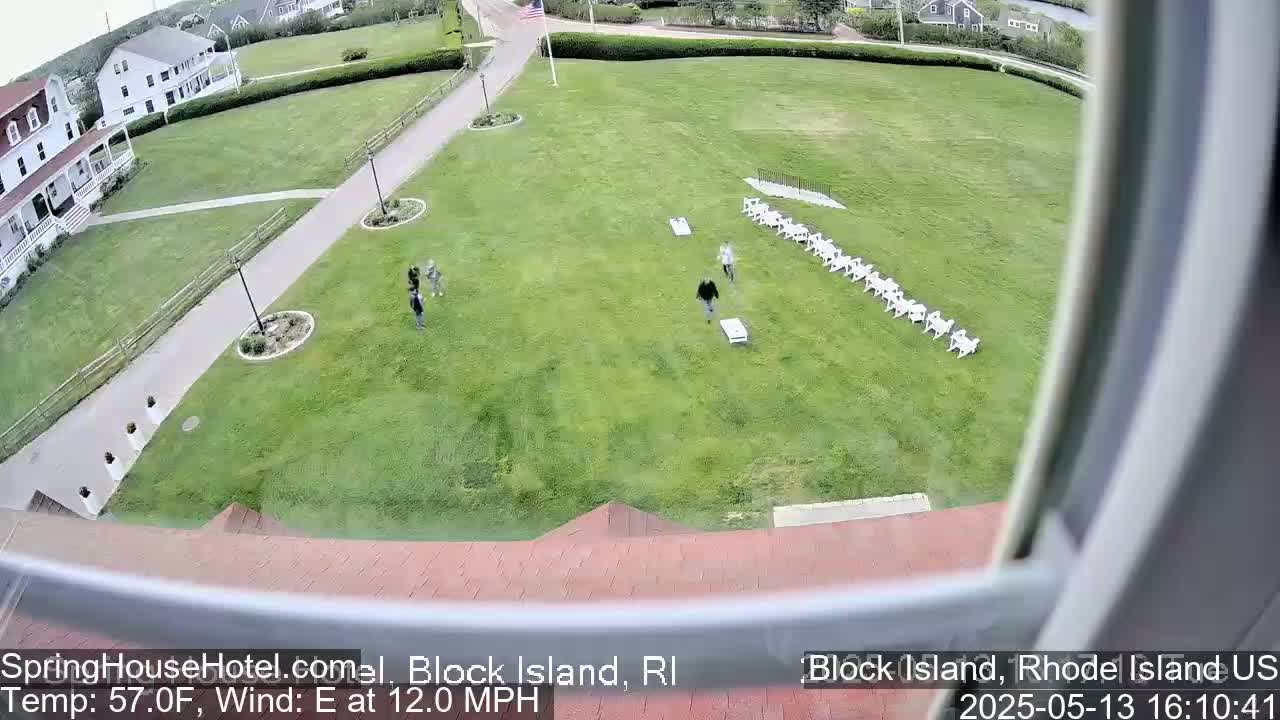 A grassy area with several people playing cornhole, a row of white chairs, and a building in the background, all under breezy conditions.