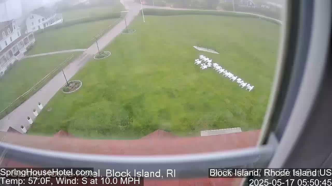 A high-angle view shows a grassy area with white tables and chairs, a building, and a walkway, all in misty conditions with a south wind.