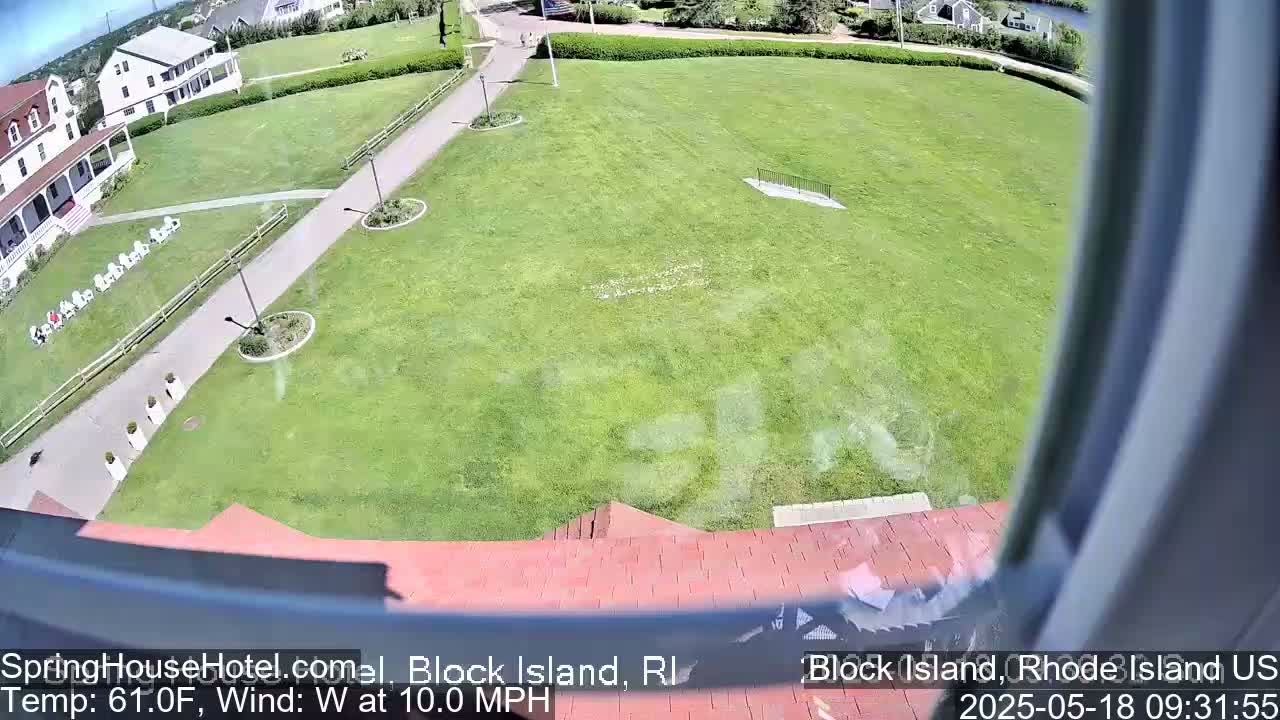 A high-angle view shows a grassy lawn, a paved walkway, and a building with white chairs on its porch, all under sunny conditions with a west wind at 10 mph.