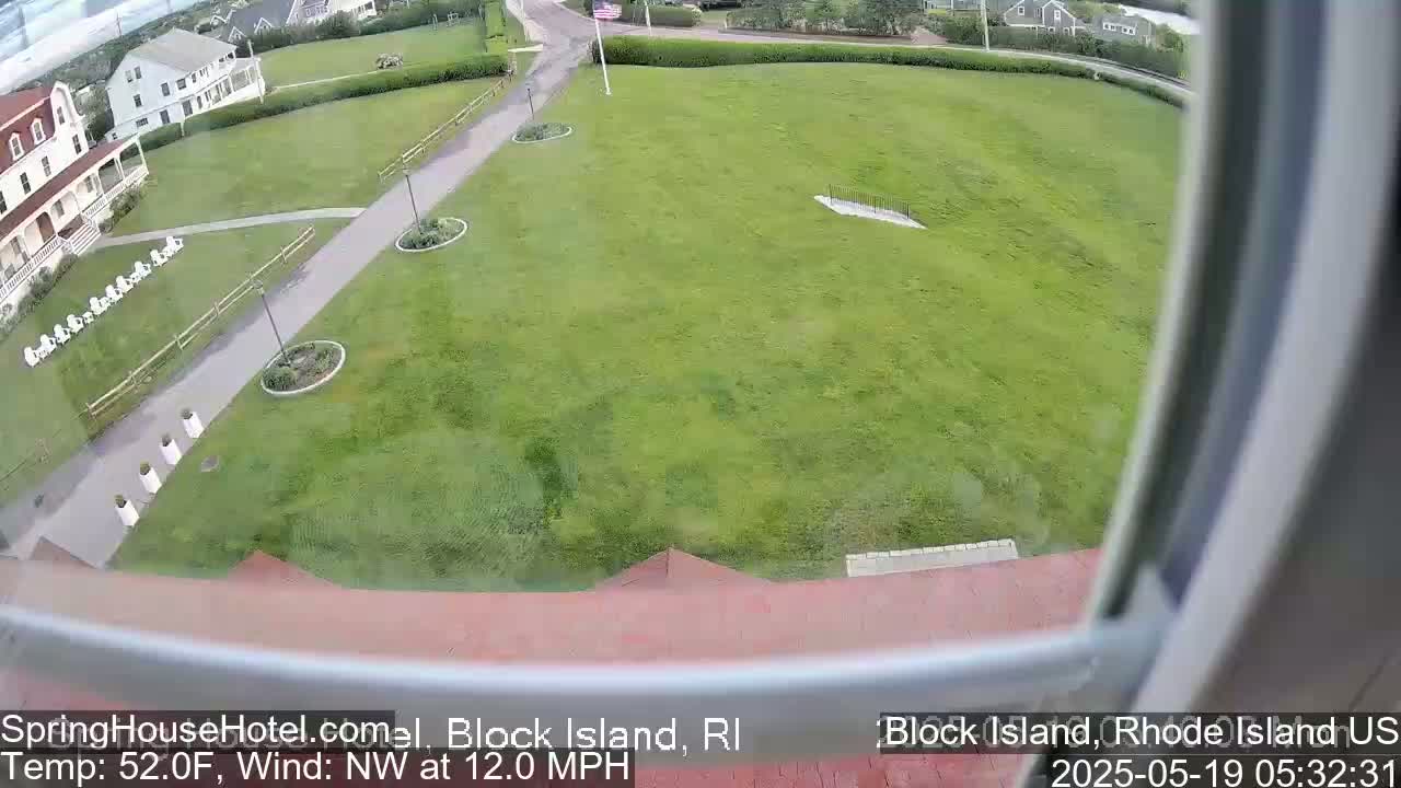 A large grassy area is seen from an elevated vantage point, with a building and a paved walkway visible in the foreground, under a partly cloudy sky with a northwest wind at 12 mph.