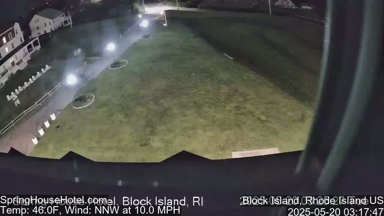A nighttime, high-angle view shows a grassy area next to a building with a paved walkway and some outdoor furniture, under a partly cloudy sky with a 10 mph northwest wind.