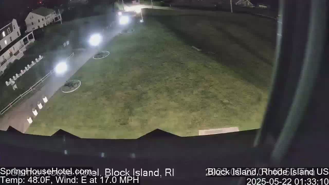 A nighttime, overhead view shows a grassy area illuminated by lights, next to a building and a paved walkway with some outdoor furniture.  The wind is blowing from the east at 17 mph.