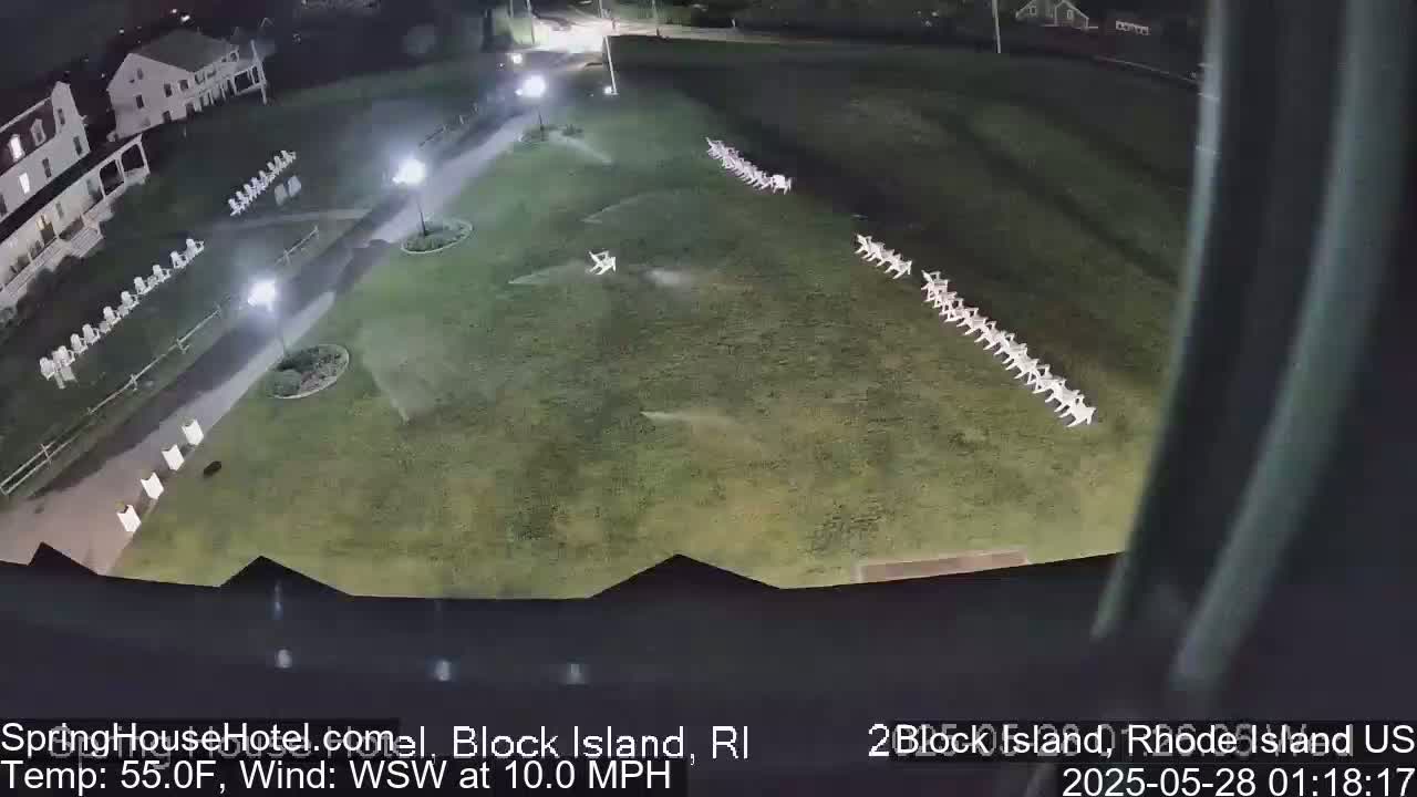 A nighttime aerial view shows rows of white chairs arranged on a grassy area next to a building, with a slight wind.