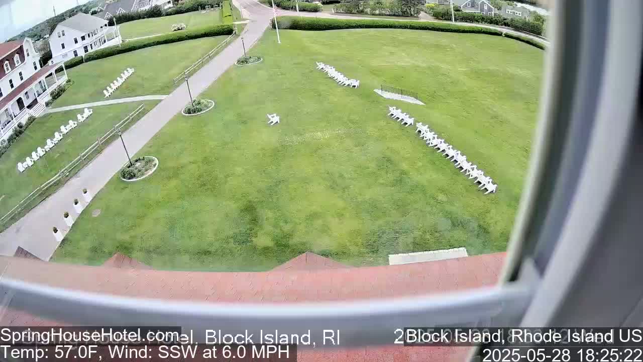 A grassy lawn with rows of white chairs is viewed from an elevated angle, with a building and pathway visible in the background on a partly sunny day with a south-southwesterly wind.