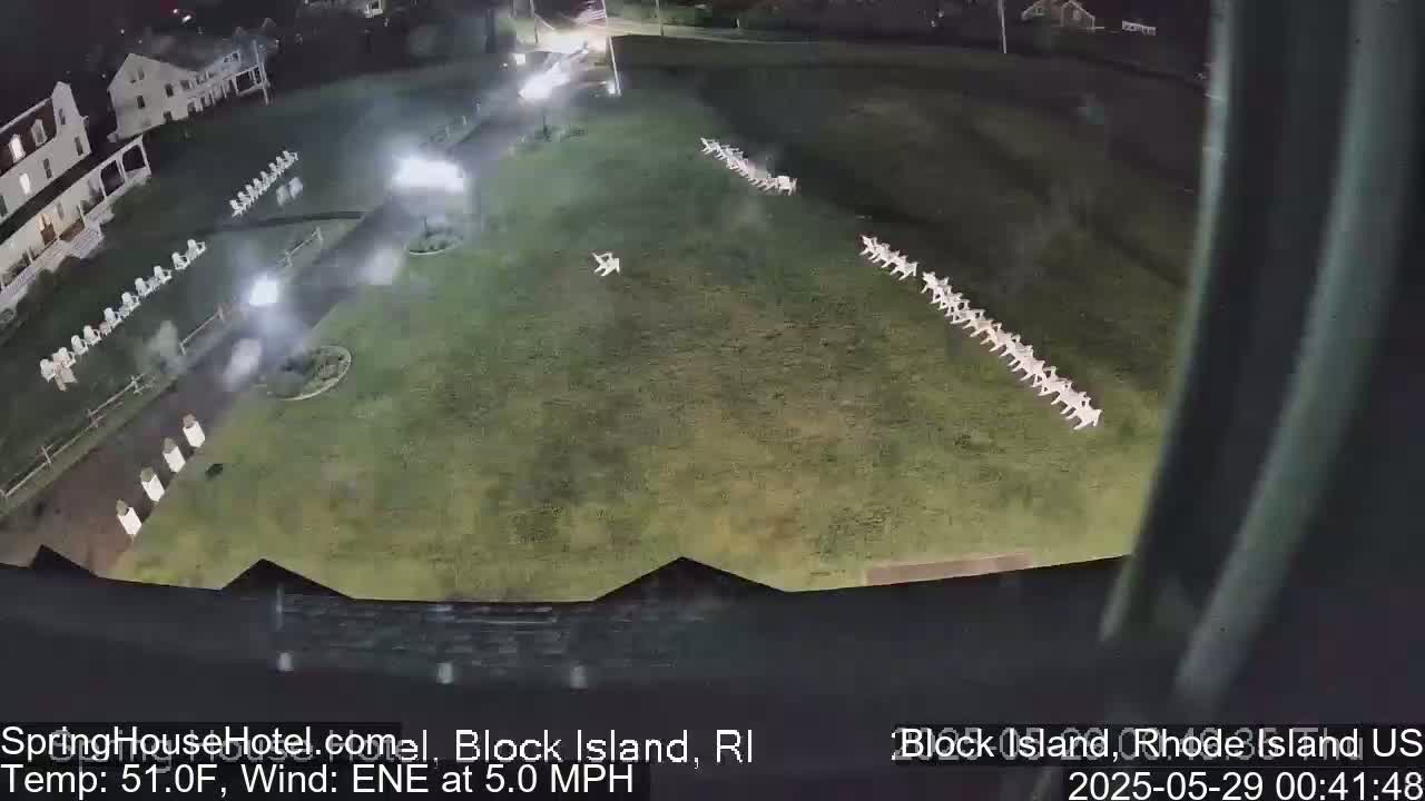 A nighttime, outdoor view shows a grassy area with rows of white chairs, a building in the background, and a slight wind.