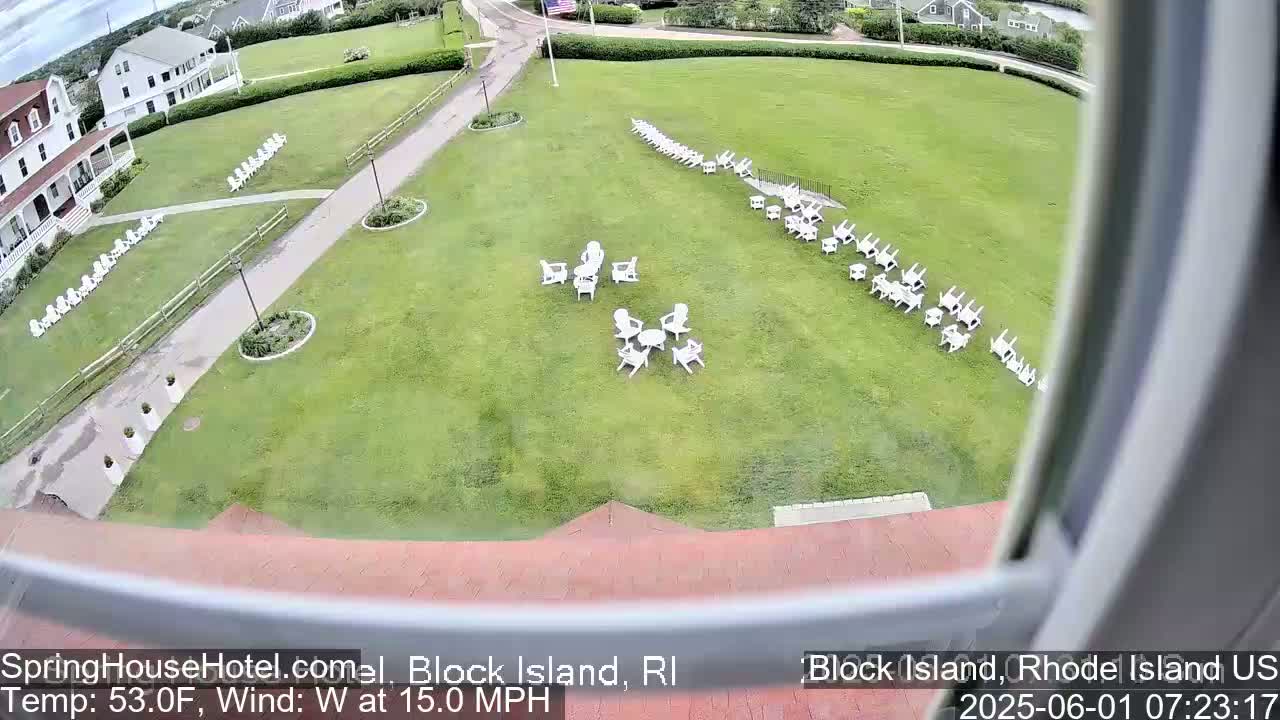 A large grassy area with numerous white chairs arranged in groups is seen from an elevated vantage point, with a building and a windy, 53°F day in the background.