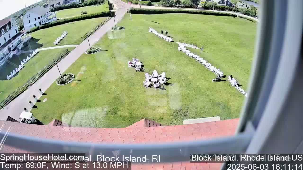 A large grassy area with white chairs arranged in rows and small groups around tables, seen from an elevated viewpoint on a sunny day with a south wind.