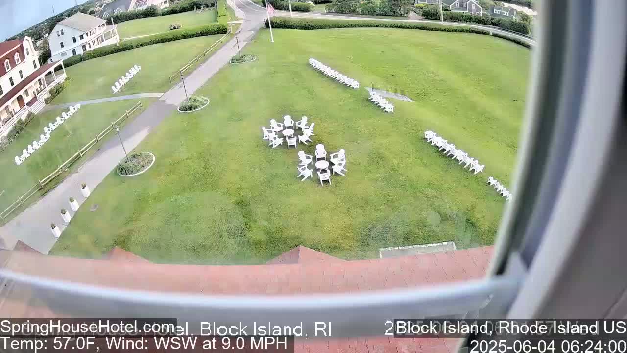 An expansive lawn with numerous white chairs arranged in rows and circular groupings surrounds a building, viewed from above on a partly sunny day with a WSW wind at 9.0 mph.