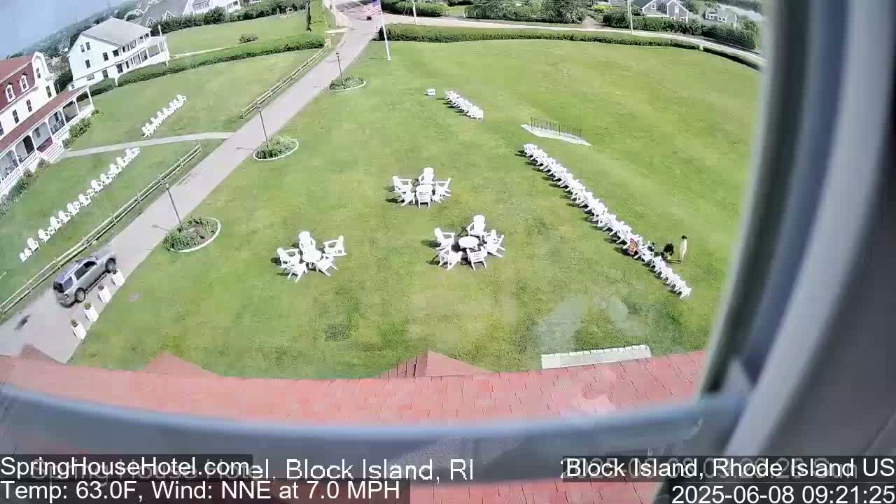 An aerial view shows a grassy area with white chairs arranged in rows and small groups around tables, a vehicle parked near a building, and two people standing near the chairs, all under a sunny sky.