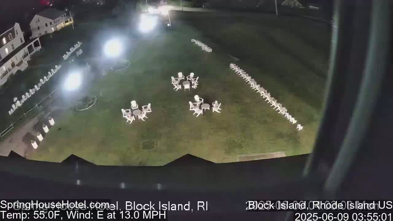 A nighttime, overhead view of a grassy area with numerous white chairs arranged in groups and lines, illuminated by bright lights, with a building visible in the background under a clear sky and a 13 mph east wind.