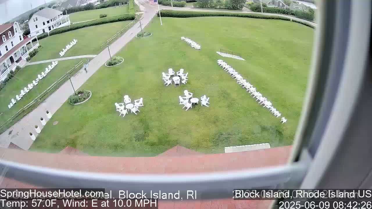 A grassy area with several sets of white chairs and tables arranged in different configurations, viewed from above on a partly sunny day with a 10 mph east wind.