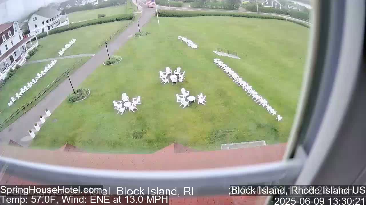 A grassy lawn with numerous white chairs arranged in groups and lines, viewed from an elevated position on a windy, overcast day.
