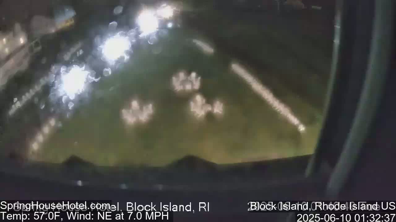 A blurry nighttime view of a dark, grassy area with faint lights and a northeast wind at 7 mph and a temperature of 57°F.