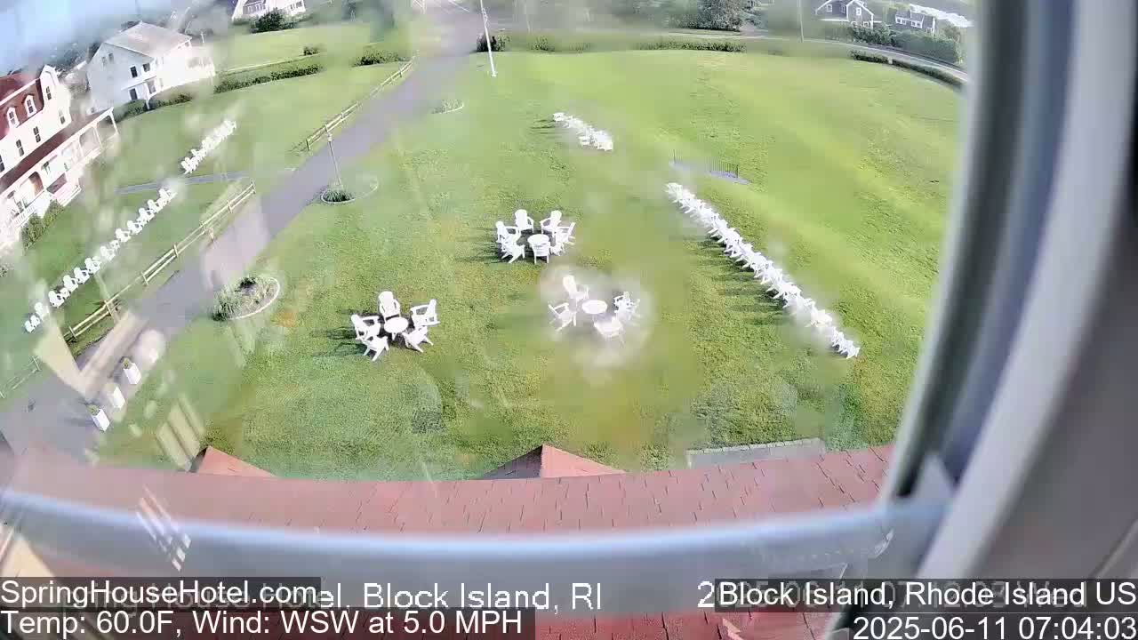 A high-angle view shows a grassy lawn with several groupings of white chairs and small tables, a building in the background, and a slightly overcast sky.
