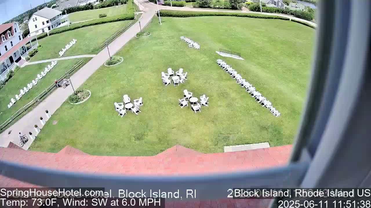 A large grassy area with numerous white chairs arranged in groups and rows, a building, and a person riding a bicycle is visible on a sunny day with a southwest wind of 6.0 mph and a temperature of 73 degrees Fahrenheit.
