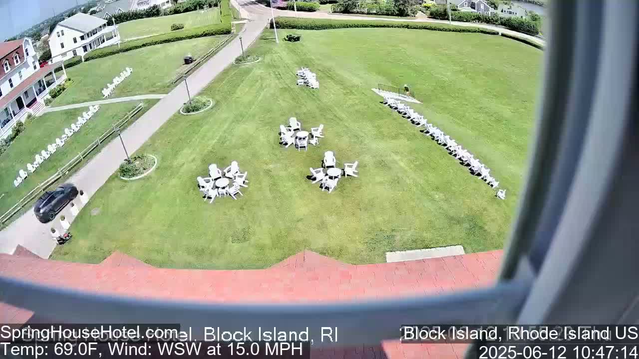 A large grassy area with several sets of white chairs and tables, a building, and a parked car is visible from a high vantage point on a sunny day with a 15 mph wind from the west-southwest.