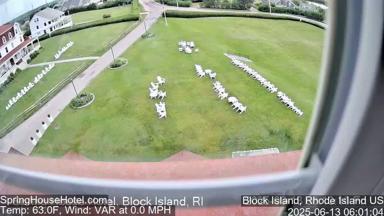 A grassy lawn with numerous white chairs arranged in rows and small groups is viewed from an elevated position on a calm day.